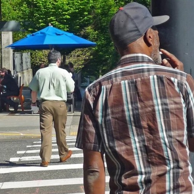 A black pedestrian waits at a crosswalk while a white pedestrian crosses ahead of him