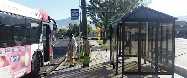 Bus riders board a bus at a stop with a shelter, sign, and benches.