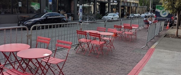 Red restaurant tables and chairs stand in the place of former curbside parking on a Seattle street