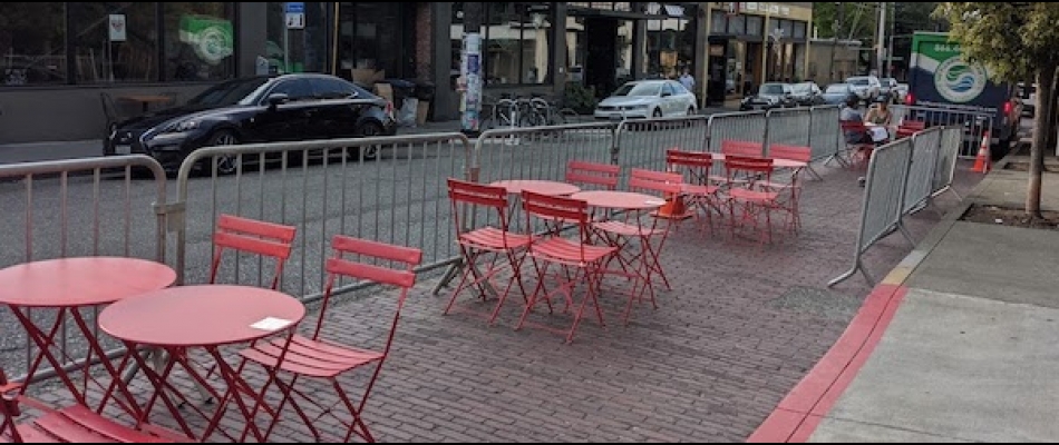 Red restaurant tables and chairs stand in the place of former curbside parking on a Seattle street